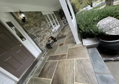 Stone porch with a bench, potted plants, and a brown door, next to a garden and driveway.