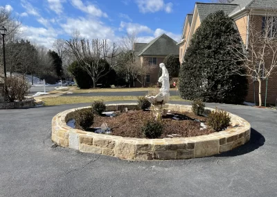 A stone-bordered circular flower bed with a white statue in the center, in front of a brick house.