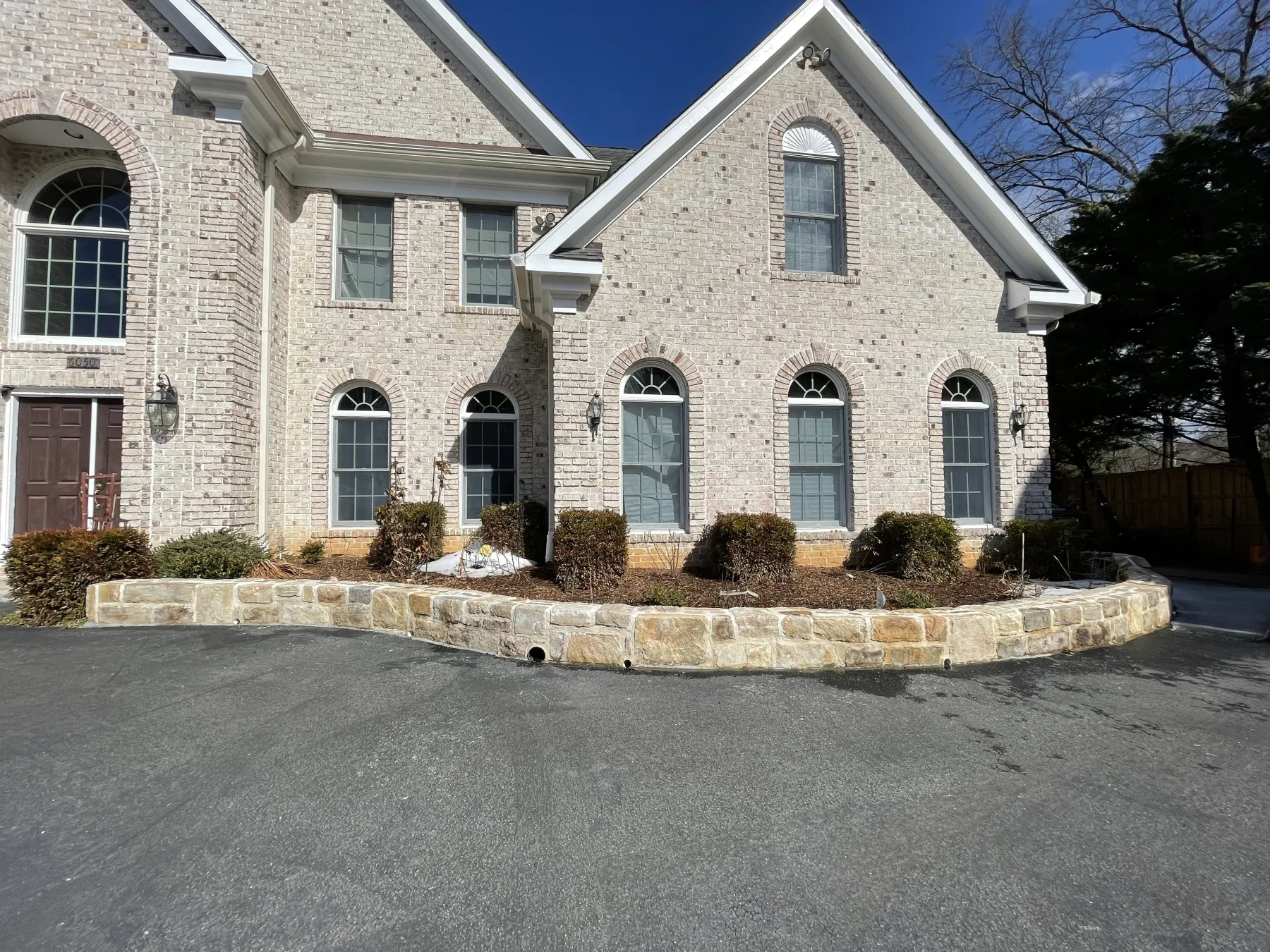 IMG_0341 Two-story light brick house with arched windows, stone-lined garden, and paved driveway on a sunny day.