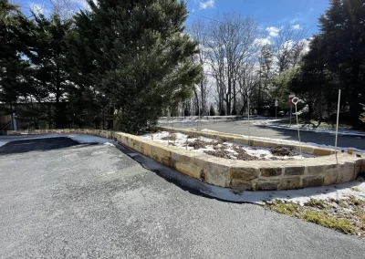 Sunlit driveway with a low stone wall and bare trees, patches of snow on the ground and planter.