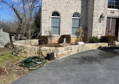 Stone raised garden bed beside a house, with bare plants, bushes, and a coiled green garden hose nearby.