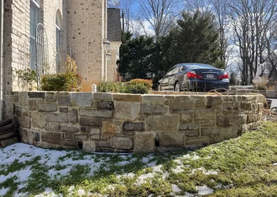 Stone retaining wall by a brick house, with a black car parked above and patches of snow on the grass below.