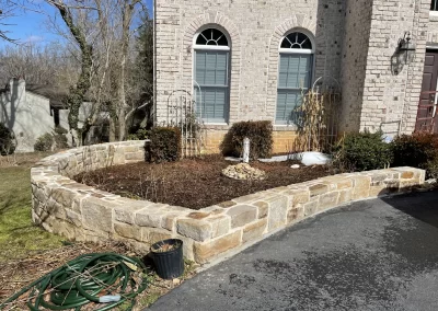 Raised stone garden bed in front of a brick house, with mulch, sparse plants, and a garden hose nearby.