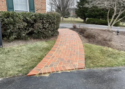 A curved brick walkway leads from a driveway to a house, bordered by shrubs and grass.