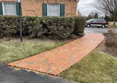 A brick pathway leads from a driveway to a house with green shutters and trimmed bushes.