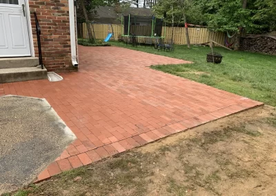 A red brick patio beside a house, with grass, a fire pit, and a trampoline in the backyard.