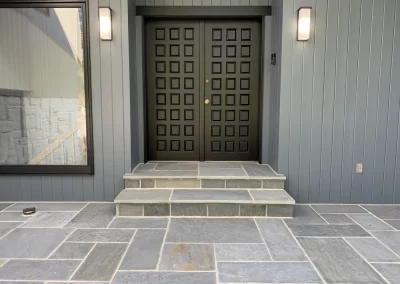 Modern double front doors with square panels, flanked by lights, and stone steps on a slate tile porch.
