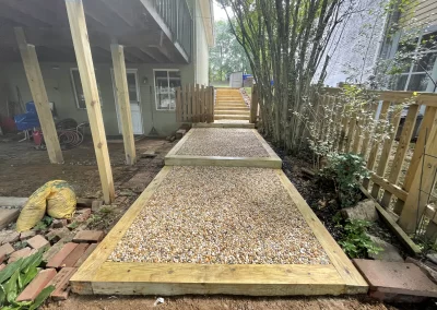 Gravel pathway with wooden borders leads up stairs between a house and a wooden fence, surrounded by plants.