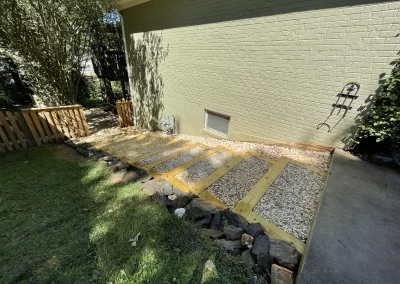 Wood and gravel steps bordered by rocks lead beside a light green brick house in a sunny yard.