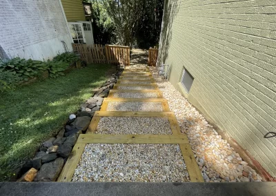 Wood-framed gravel steps lead down between two houses toward a wooden gate and a shaded backyard.