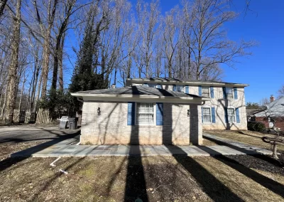 A beige brick house with blue shutters and bare trees casting shadows on a sunny day.