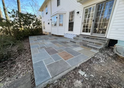 A stone patio with steps leading to a white house, surrounded by dirt and some trees in the background.