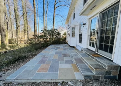 A stone patio with steps leads to a white house, surrounded by trees and greenery on a sunny day.