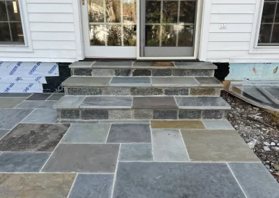 Stone steps and patio leading to a glass door, with a white exterior wall and some exposed foundation.