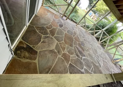 Natural stone patio with irregular shapes, next to a house and facing a backyard garden with greenery.