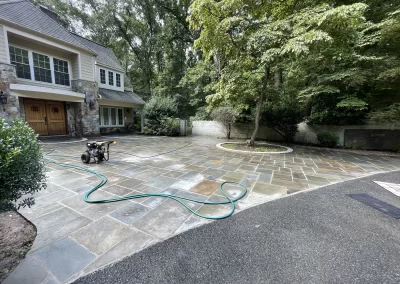 Stone driveway with a hose and pressure washer in front of a house, surrounded by trees and greenery.