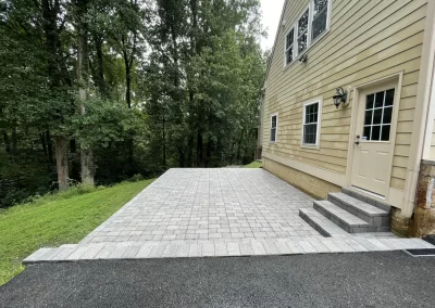 Light gray stone patio with steps beside a tan house, bordered by grass and trees.