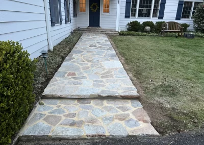 Stone walkway leading to a blue front door of a white house with dark shutters and bushes on either side.