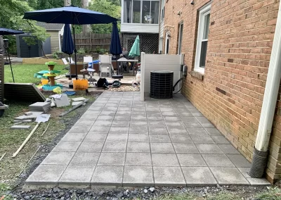 Freshly laid gray patio pavers beside a brick house, with tools and patio furniture in the background.