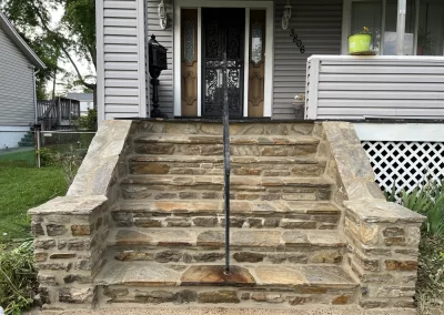 Stone steps with a black metal railing lead to a house entrance with double doors and light gray siding.