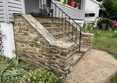 Stone front steps with a black metal railing, leading up to a house with gray siding and plants nearby.