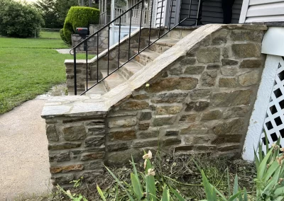 Stone steps with black railings lead to a porch, with green grass and plants in the foreground.