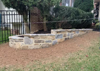 Stone retaining wall with fresh soil in front, set against shrubs and a black metal fence in a yard.