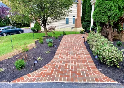 Red brick walkway with black edging, lined with plants and small path lights, leading to a house entrance.
