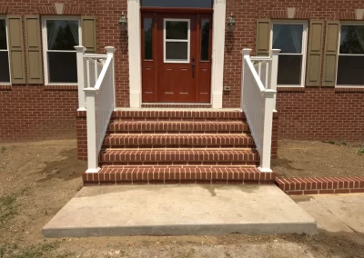 Brick steps with white railings lead to a red front door of a brick house with tan window shutters.
