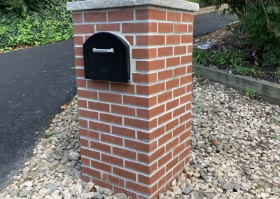 A black mailbox is mounted on a brick pillar surrounded by rocks and greenery.