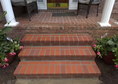 Three brick steps lead up to a porch with two chairs and potted plants on either side.