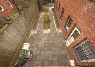 A rectangular brick patio with pavers, a small tree, and garden supplies near a red brick building.