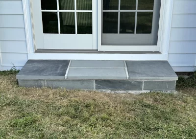 Gray stone step in front of glass double doors, surrounded by grass and light-colored siding.