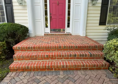 Red brick steps leading to a pink front door with white trim, flanked by bushes and windows.