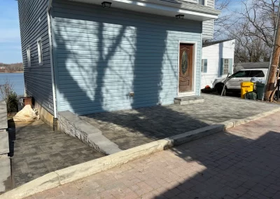 A house with new gray concrete paving and a wooden door, next to a car and trash bins on a sunny day.