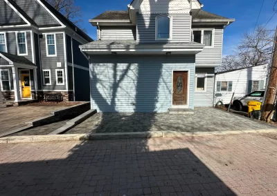A gray house with a wooden door, driveway, and shadows of tree branches on the ground.