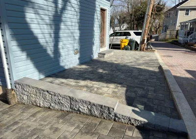 A paved driveway with a stone retaining wall beside a blue house, a white car, and trash bins in the background.