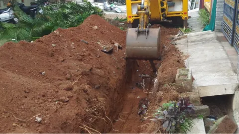 A yellow excavator digging a trench next to a sidewalk and some plants.